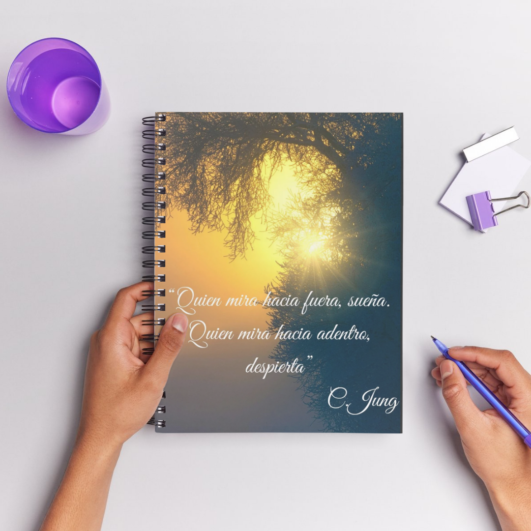 Una mano sostiene un bolígrafo ante una agenda en cuya portada vemos el sol atravesando las ramas de los árboles con el texto "Quien mira hacia fuera, sueña; quien mira hacia adentro, despierta" C. Jung.